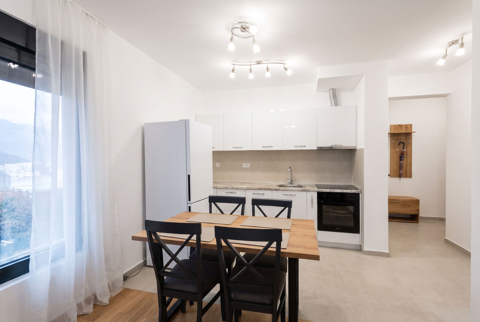 Modern kitchen with white cabinets, wooden dining table, black chairs, and refrigerator near the window.