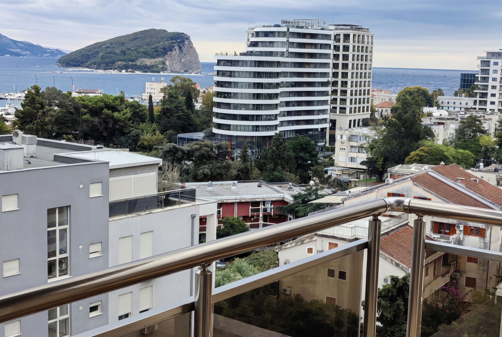 View from a balcony overlooking the city, sea, and island. Metal railing with glass panels in foreground.