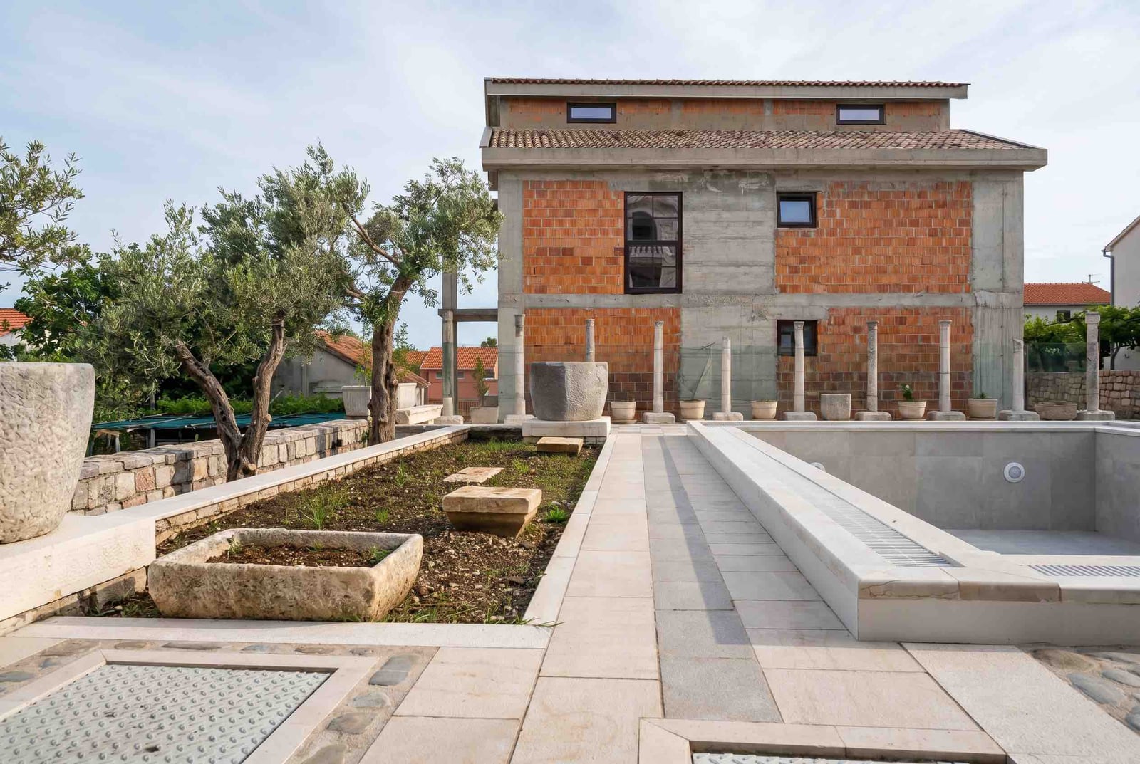 House under construction in Luštica, Montenegro: brick facade, swimming pool, stone columns, and landscaped grounds.