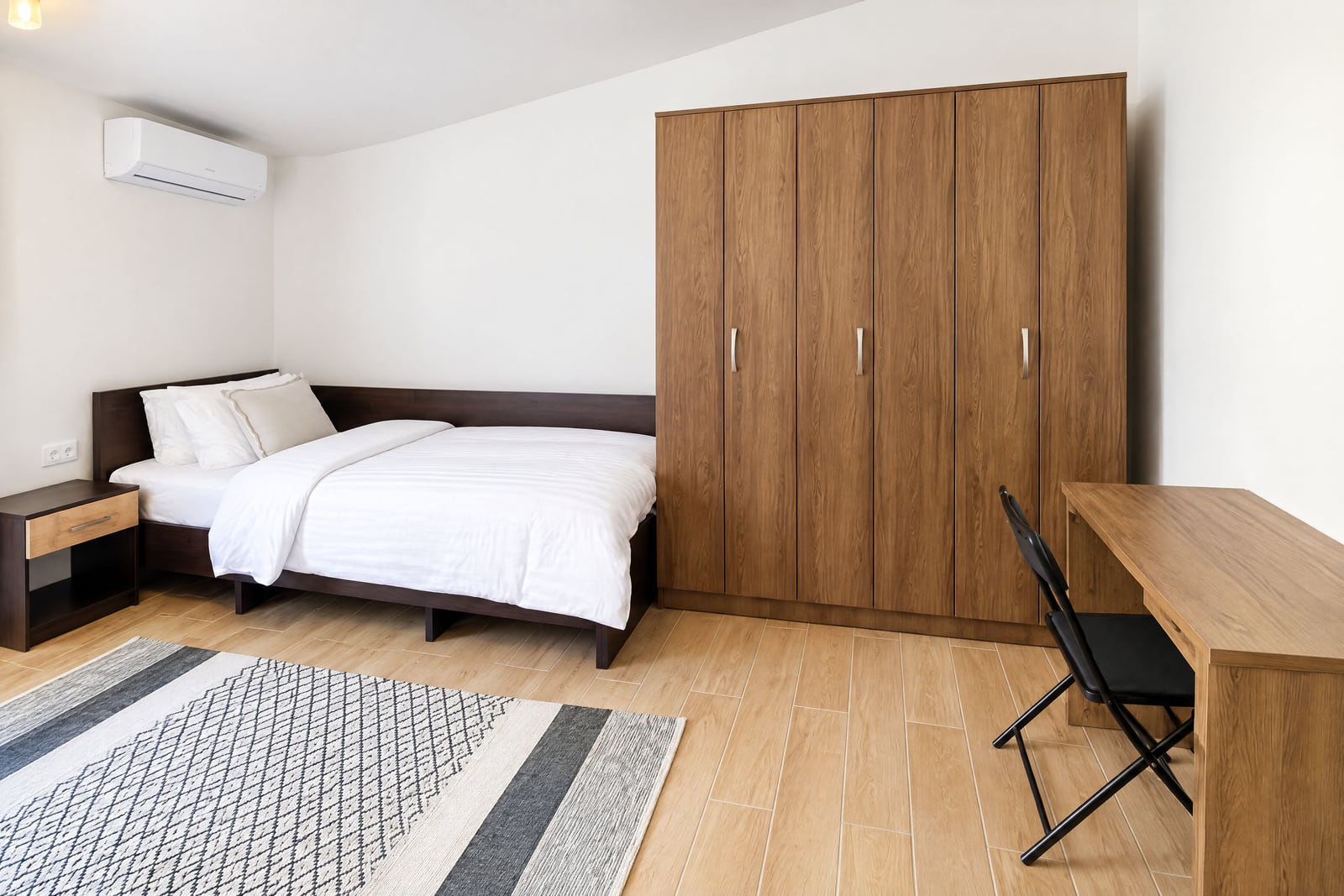 Bedroom featuring a double bed, large wooden wardrobe, desk with chair, and wall-mounted air conditioner.