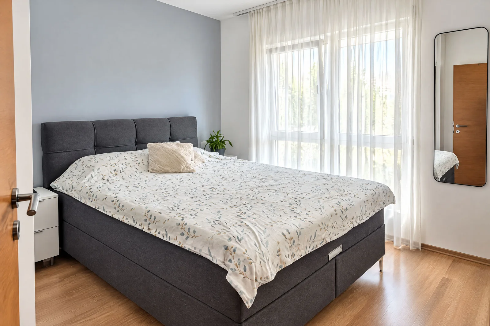 Modern bedroom featuring a grey upholstered bed with floral bedding, sheer curtains, and a wall mirror.
