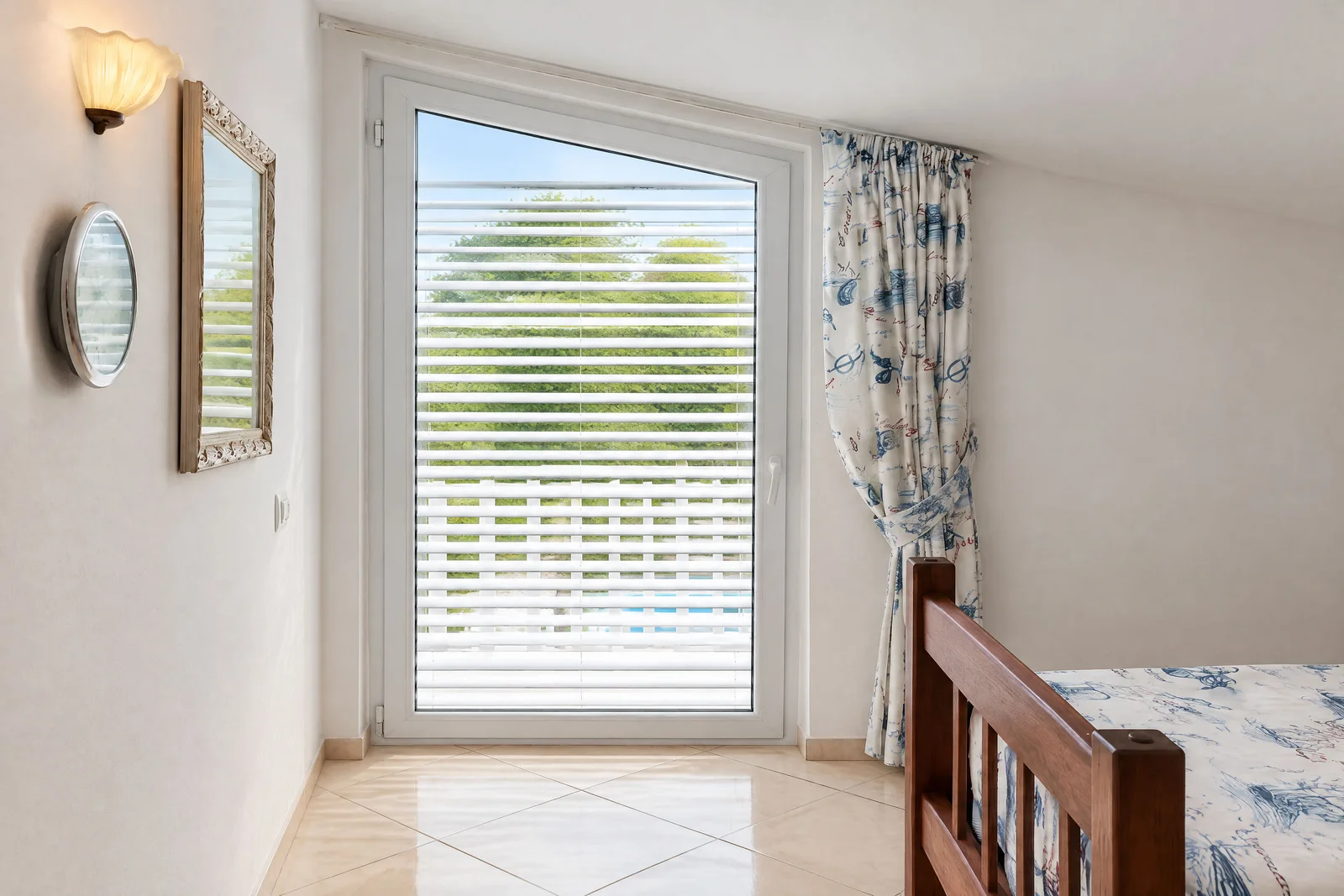 Bedroom with a wooden bed frame, wall mirror, and sconce next to a window with blinds.