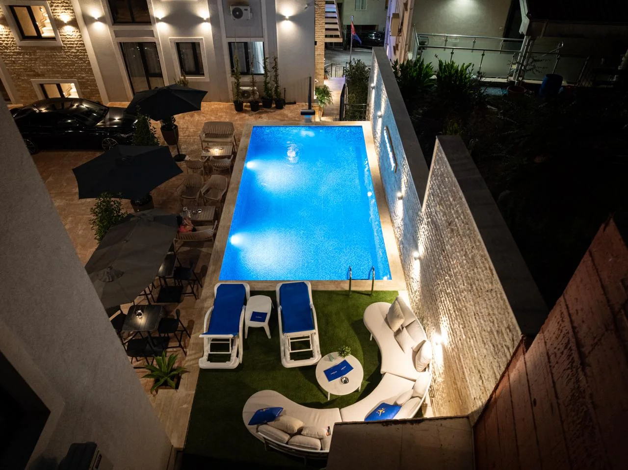 Aerial night view of an illuminated blue swimming pool surrounded by lounge chairs and umbrellas.