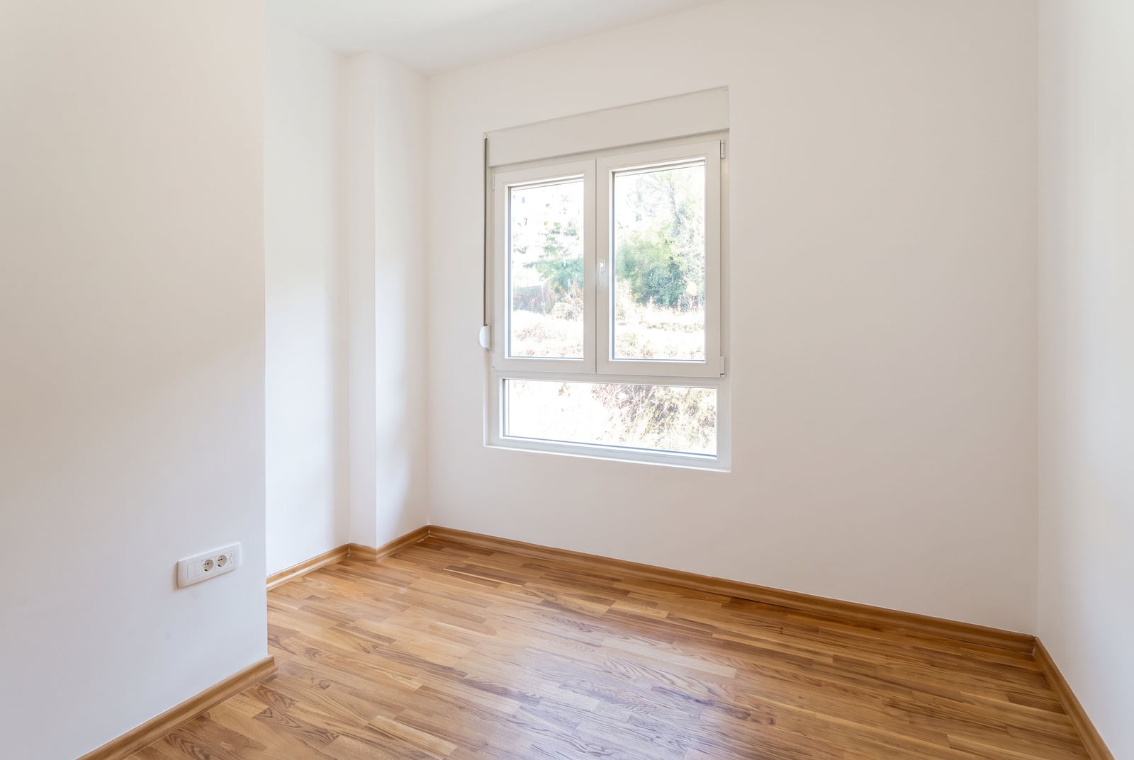 Bright empty bedroom with white walls, parquet flooring, and a window in an apartment in Budva, Montenegro.