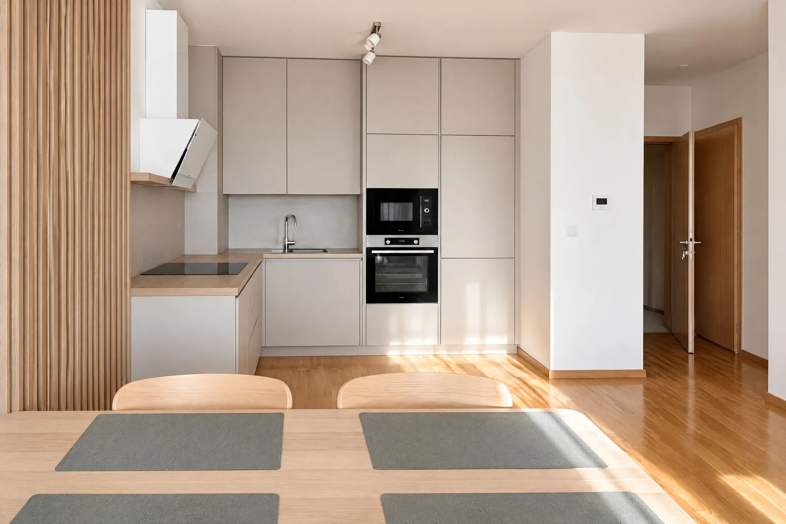 Modern kitchen with light cabinets, built-in oven, microwave, sink, and a wooden dining table with placemats in the foreground.