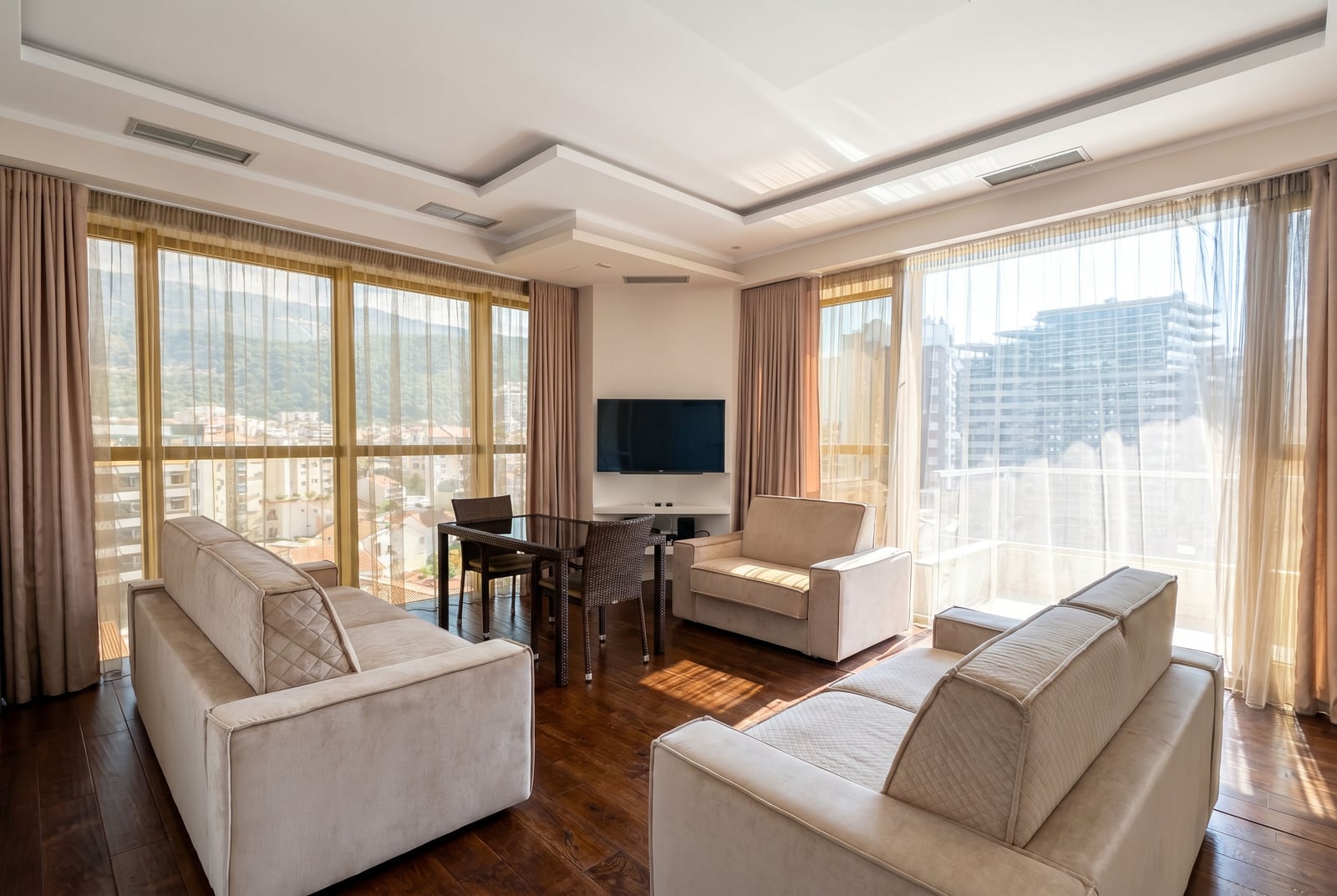 Bright living room in a Budva apartment, Montenegro: upholstered furniture, dining area, and floor-to-ceiling windows with a city view.