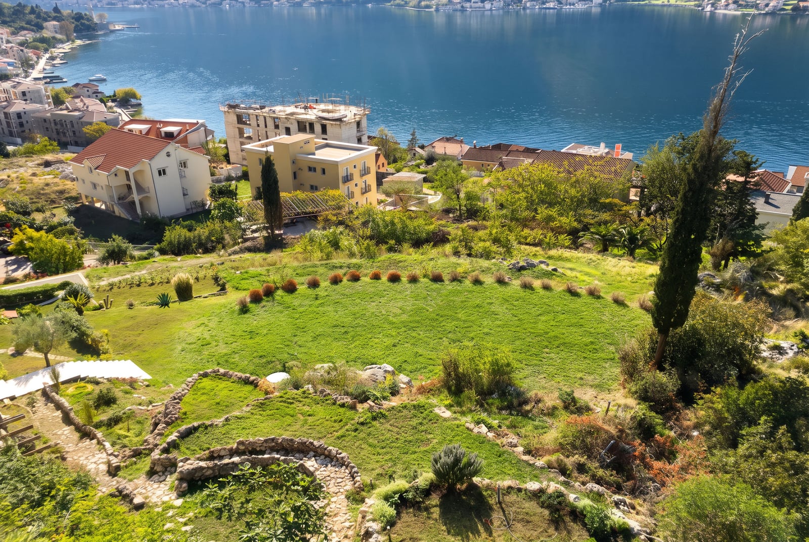 View of a green slope with stone terraces, houses, and a large body of water in the background.