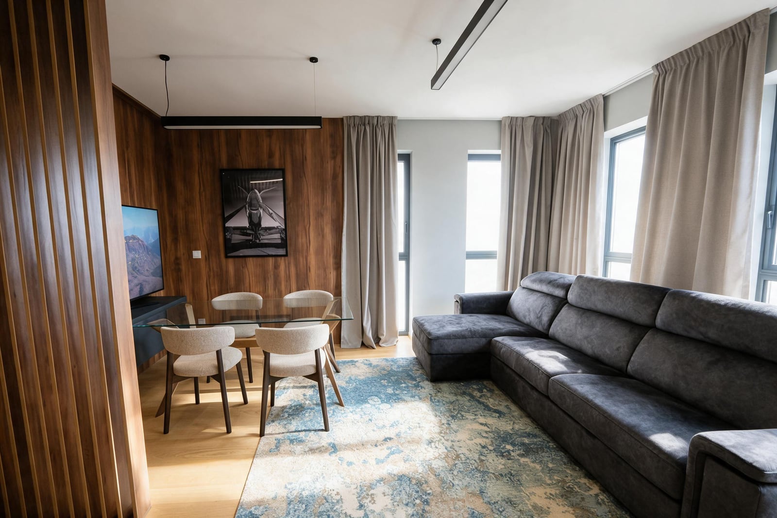 Modern living room featuring a grey sectional sofa, glass dining table, wood wall paneling, and TV.