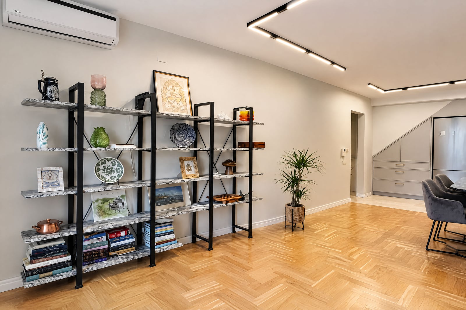 Living room with parquet flooring, open shelving unit with decor and books, potted plant, and dining area edge.