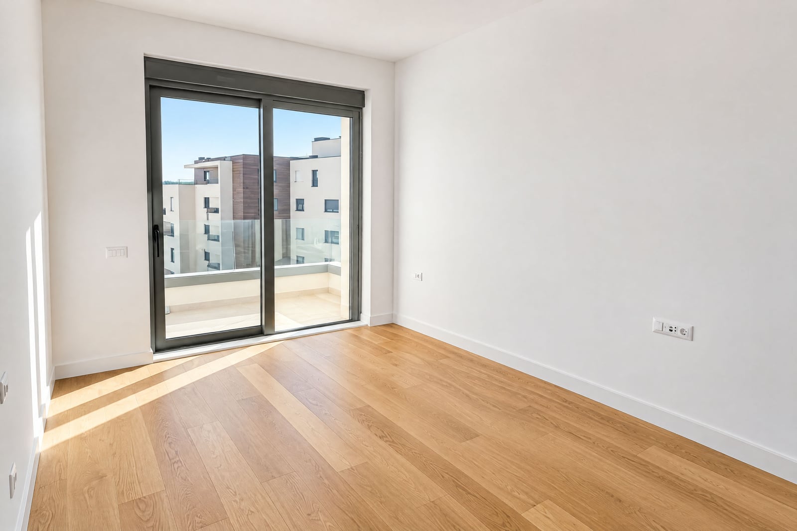 Empty room with light wood flooring and white walls, featuring a large sliding glass door leading to a balcony.