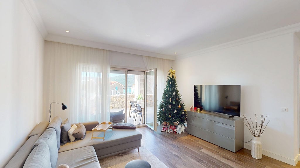 Bright living room with terrace access in Luštica Bay, Montenegro: gray sofa, TV area, and a Christmas tree.