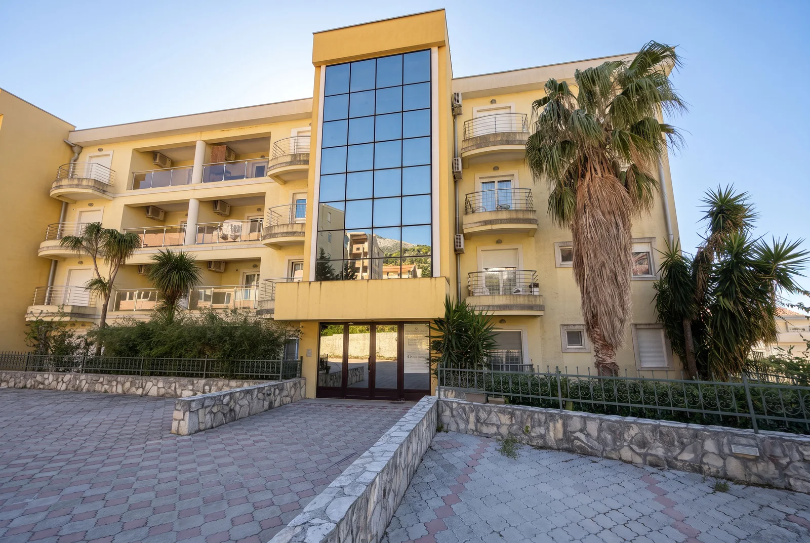 View of a yellow multi-story apartment building with balconies, palm trees, and stone fence.