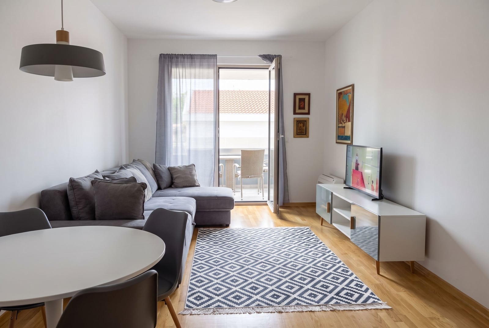 Bright room with a grey sofa, white table, TV on a stand, and a patterned rug on wooden flooring.