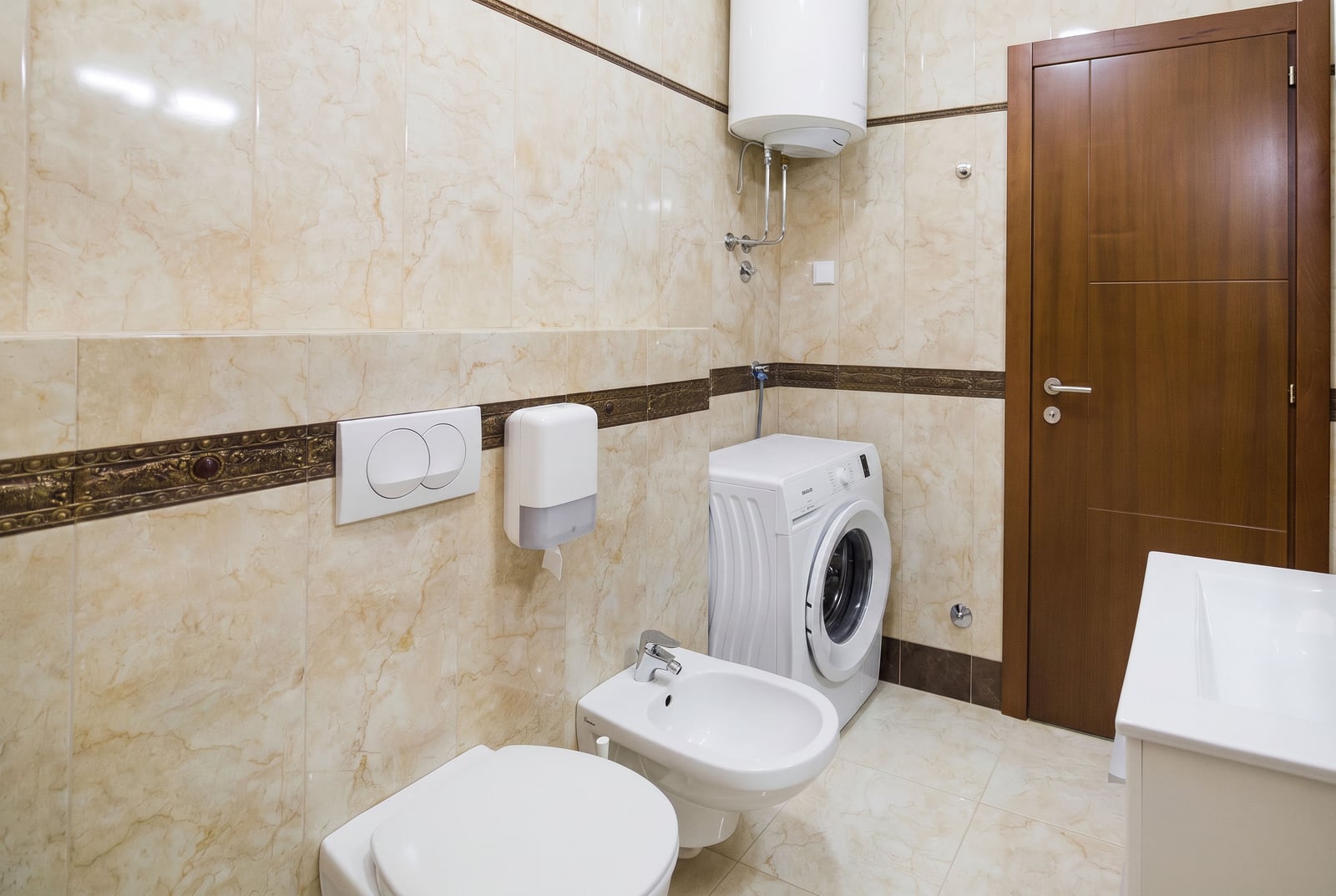 Bathroom in an apartment in Becici, Montenegro: beige tiles, wall-mounted toilet, bidet, washing machine, and water heater.