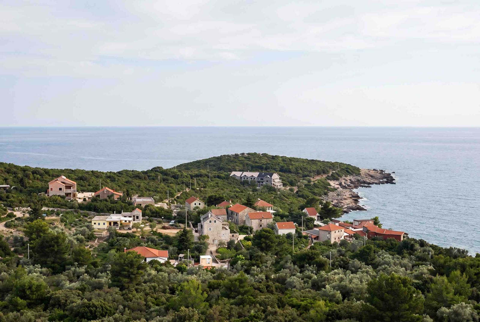 Panoramic view of stone houses in Luštica, Montenegro, surrounded by greenery and the Adriatic Sea coastline.