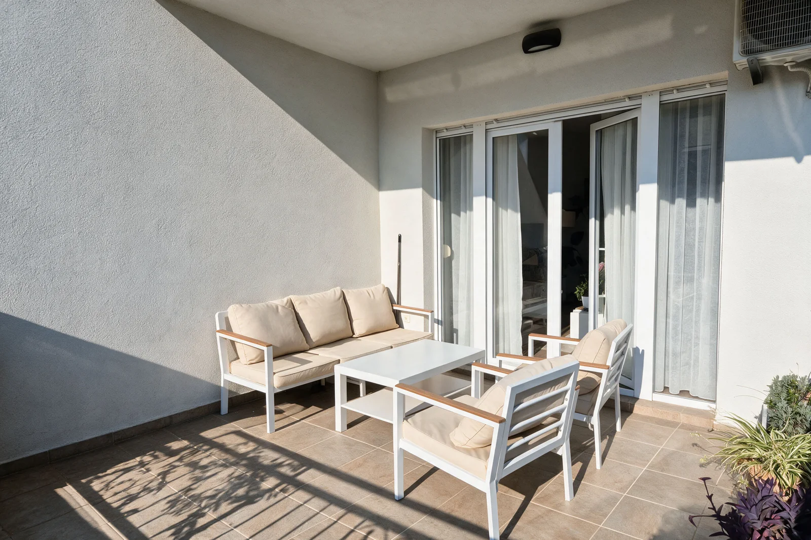 Balcony with white outdoor furniture and beige cushions next to sliding glass doors on tiled floor.