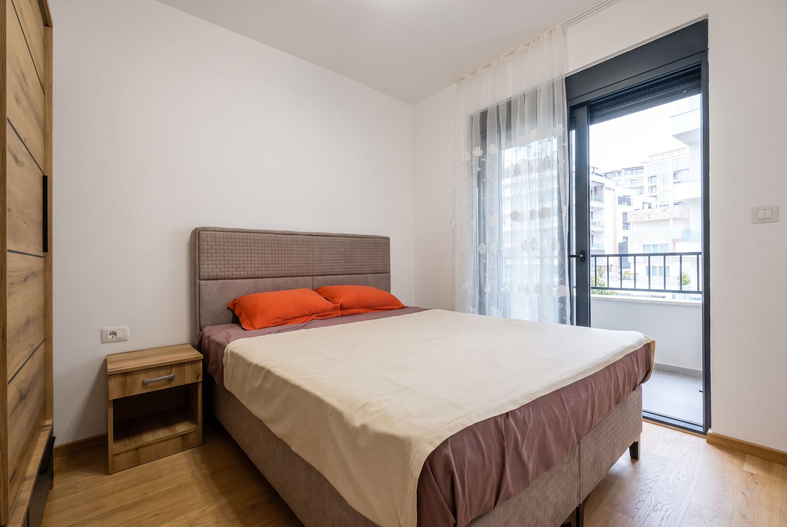 Bedroom featuring a double bed with orange pillows, a wooden nightstand, wardrobe, and balcony access.