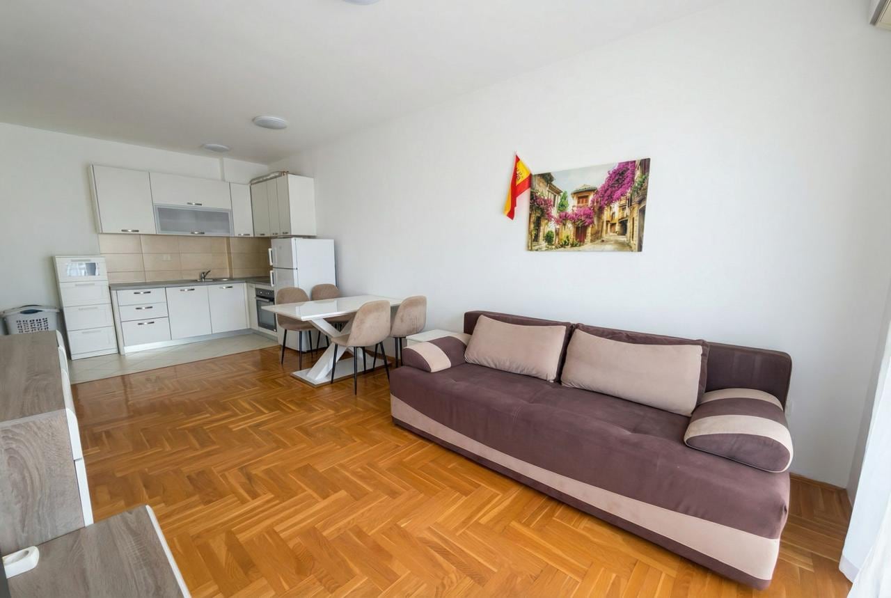 Living room with kitchen in an apartment in Budva, Montenegro: brown sofa, dining area, and light-colored kitchen set.