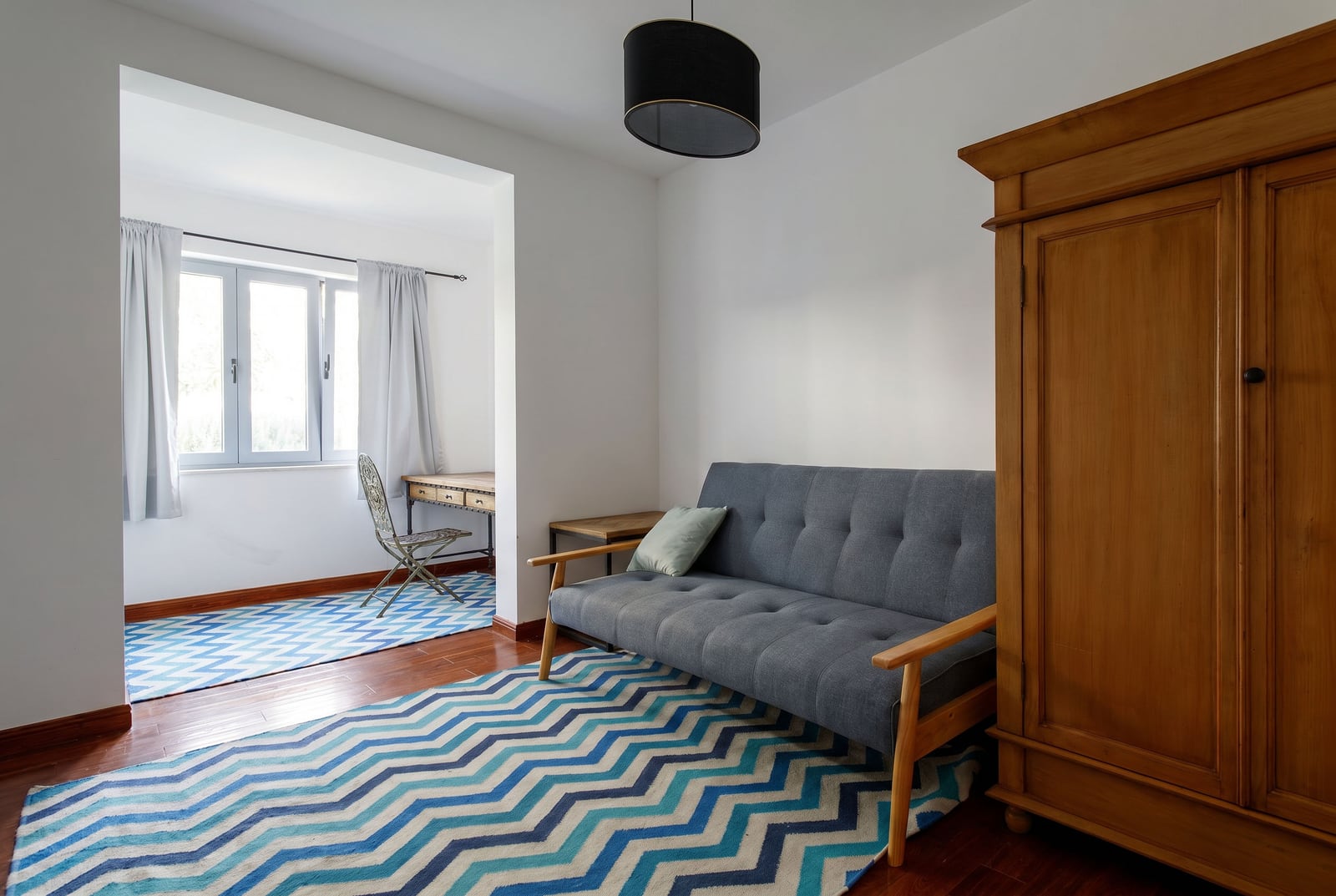 Living room in an apartment in Risan, Montenegro: gray sofa, wooden wardrobe, workspace by the window, and zigzag-patterned rugs.