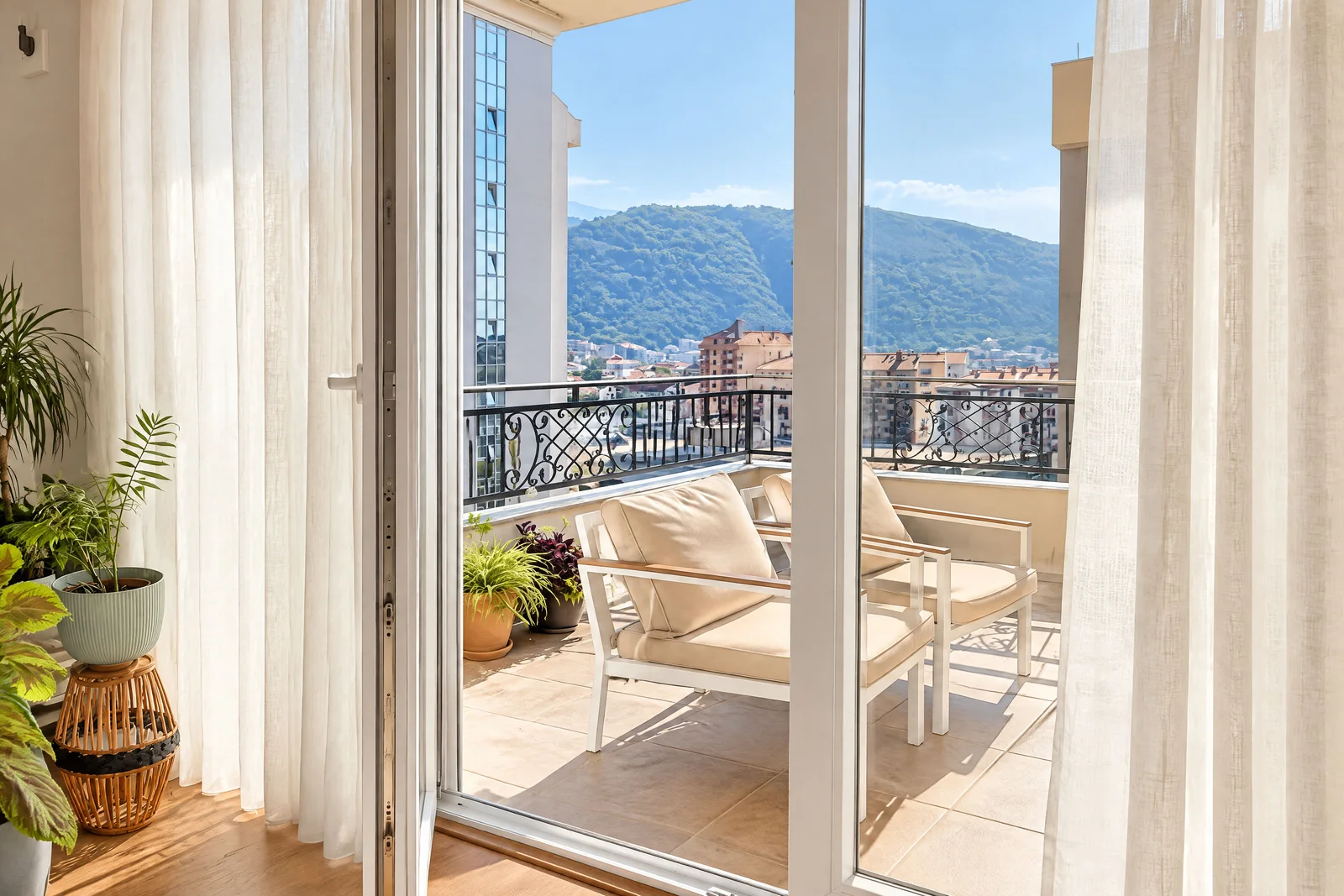 View of a balcony with white chairs, potted plants, and a cityscape with mountains in the background.