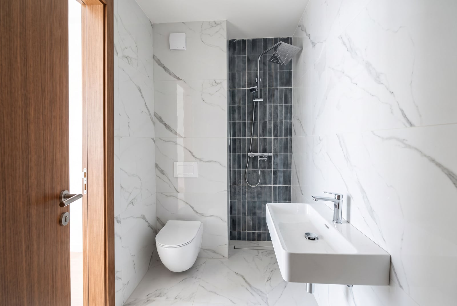 Modern bathroom with white marble tiles, wall-mounted toilet, rectangular sink, and shower area with dark vertical tiles.
