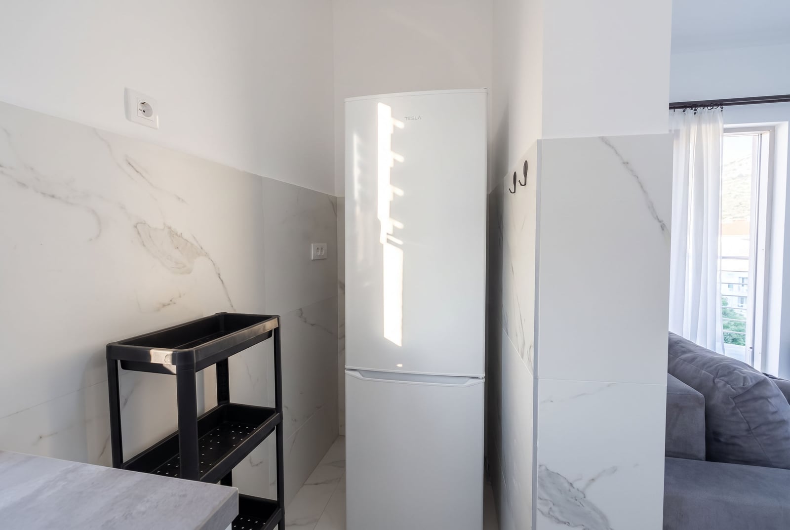 White refrigerator and black shelving unit against marble-effect tiles; part of a grey sofa is visible nearby.