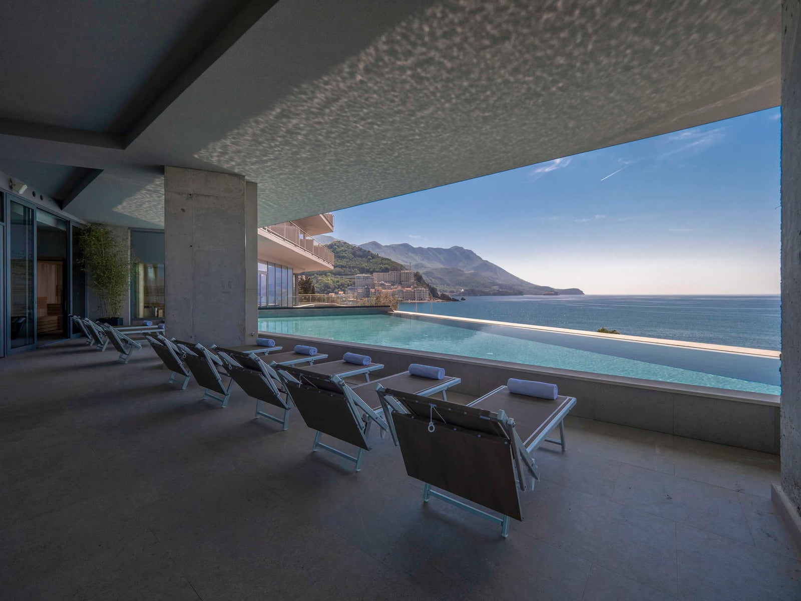 Infinity pool with lounge chairs under a covered terrace overlooking the sea and mountains.
