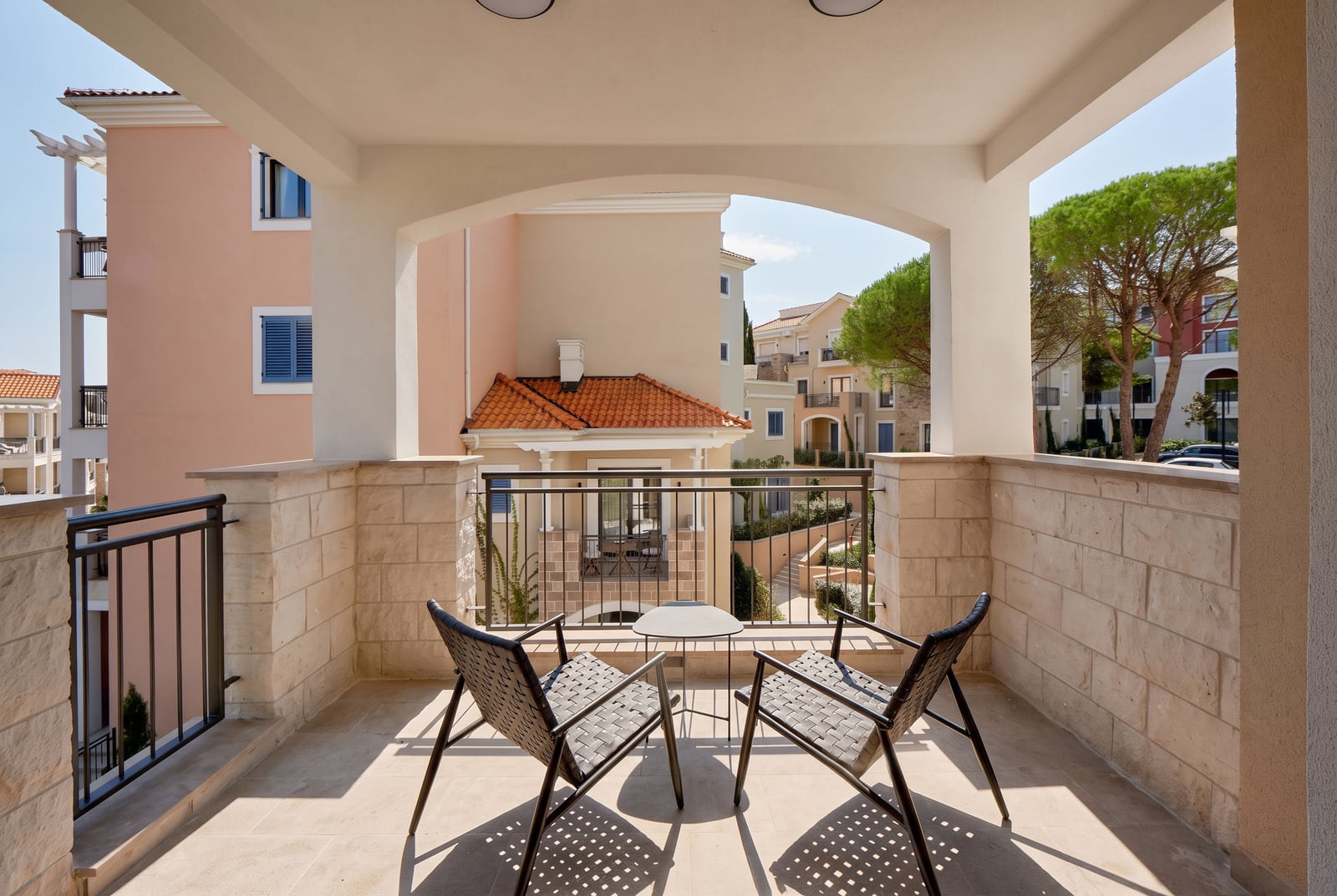 Covered balcony with stone walls, two woven chairs and a small table overlooking residential buildings.