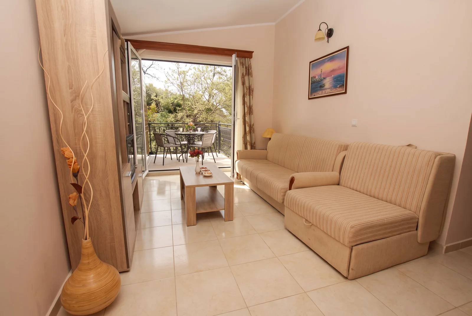 Living room with a beige sofa, wooden coffee table, and open doors leading to a balcony overlooking trees.