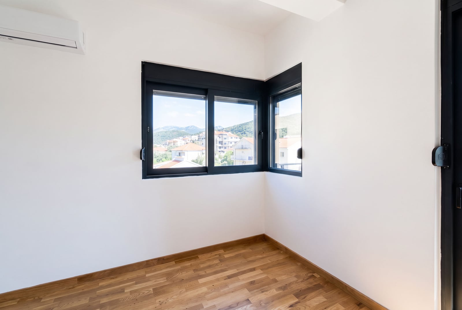 White bedroom with parquet flooring, air conditioning, and a corner window overlooking the city and mountains in Bar, Montenegro.