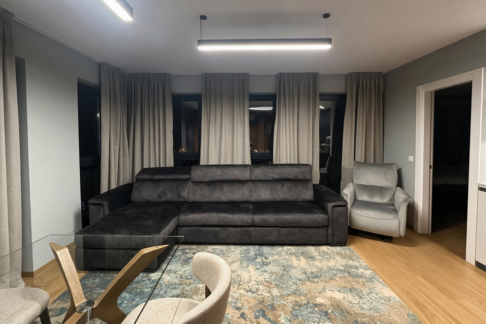 Modern living room featuring a dark gray sectional sofa, light armchair, glass table, patterned rug, and wood flooring.