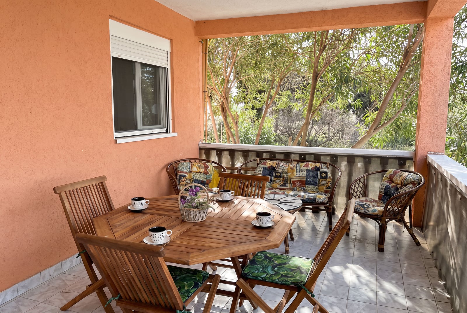 Balcony with wooden dining table, wicker furniture, and view of trees. Orange walls and tiled floor.