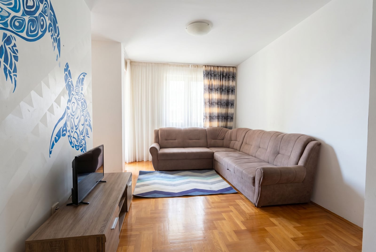 Living room in an apartment in Bečići, Budva, Montenegro: corner sofa, TV stand, parquet flooring, and turtle wall decor.