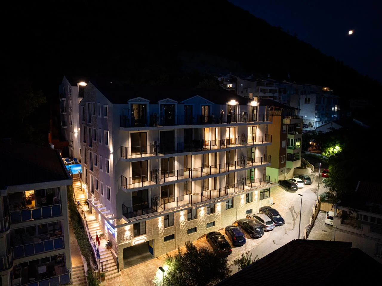 Night exterior view of a multi-story hotel building with balconies, illuminated facade and parking lot.