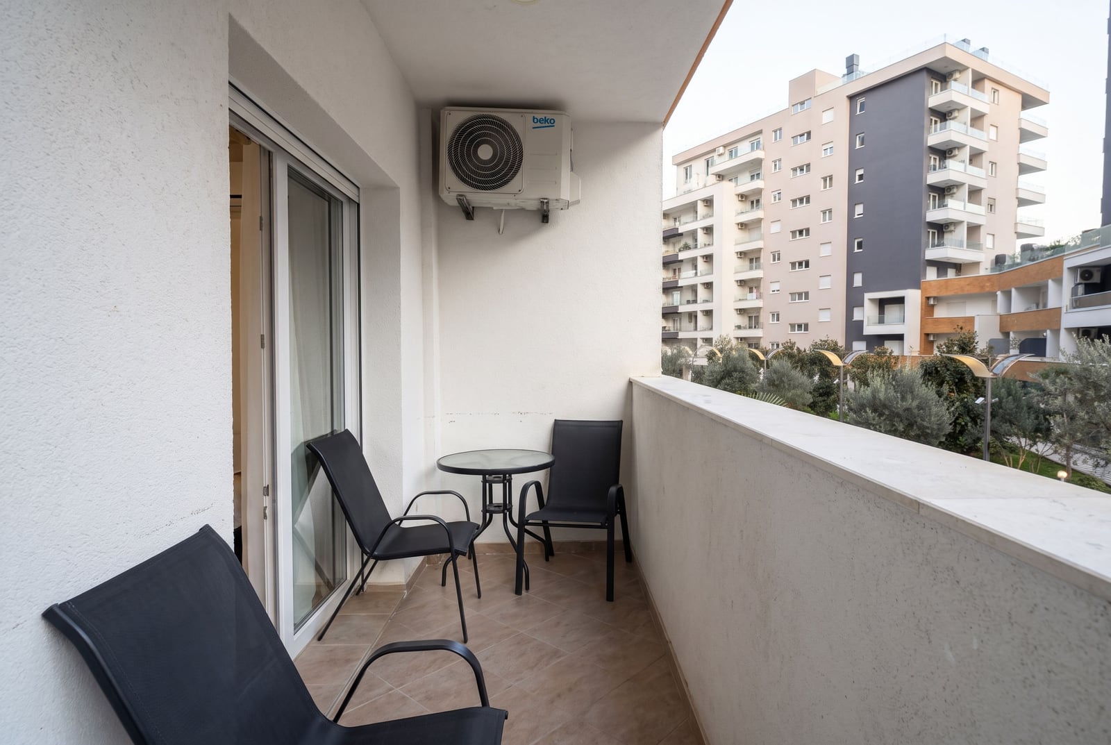 Balcony with black chairs, small table, wall-mounted AC unit, and view of apartment buildings.