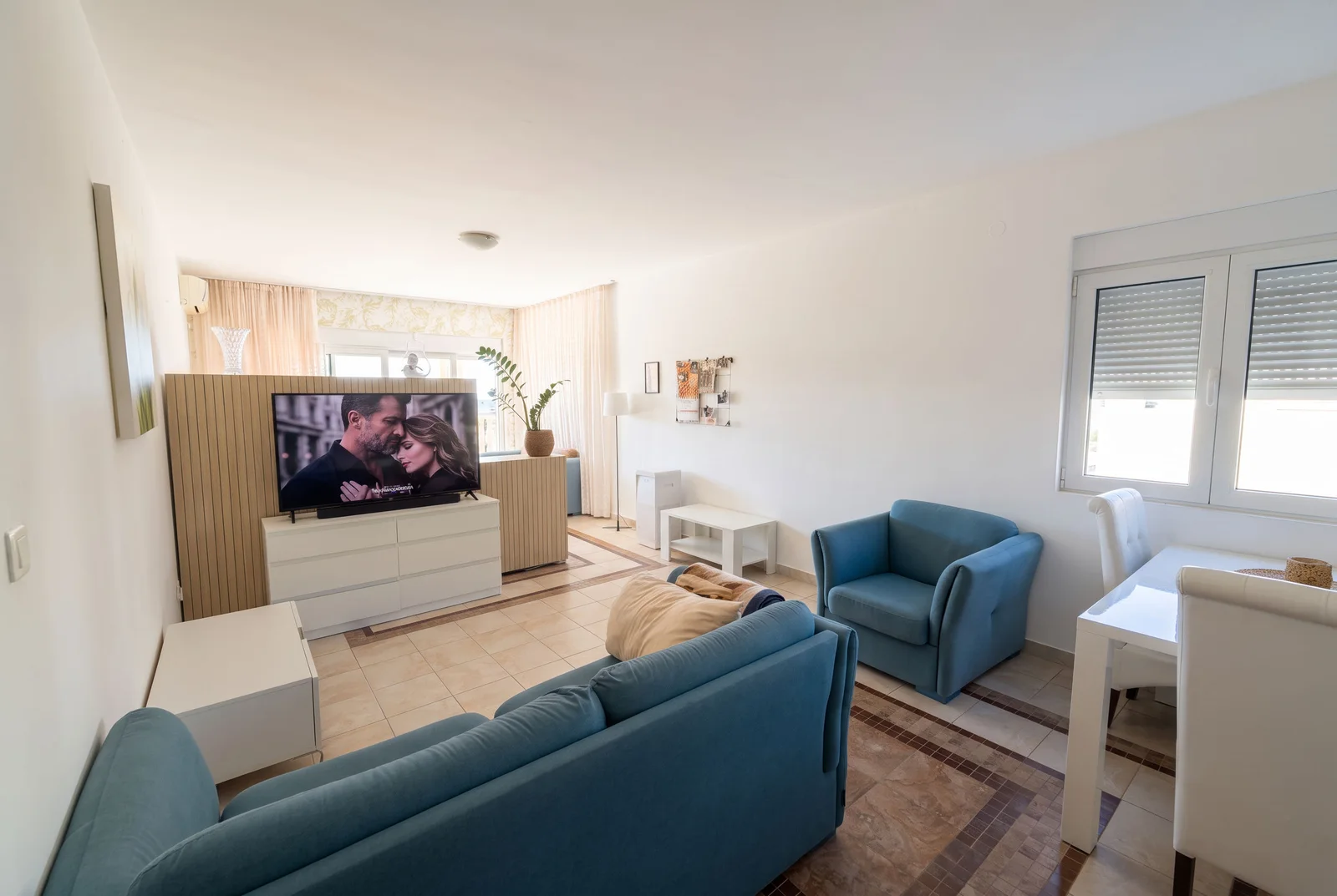 Bright room with a blue sofa, armchair, TV on a white cabinet, and a white dining table near the window.