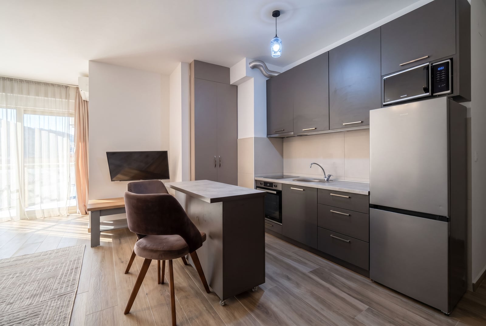 Modern kitchen with grey cabinets, fridge, microwave, and a bar counter with chairs next to a living area.