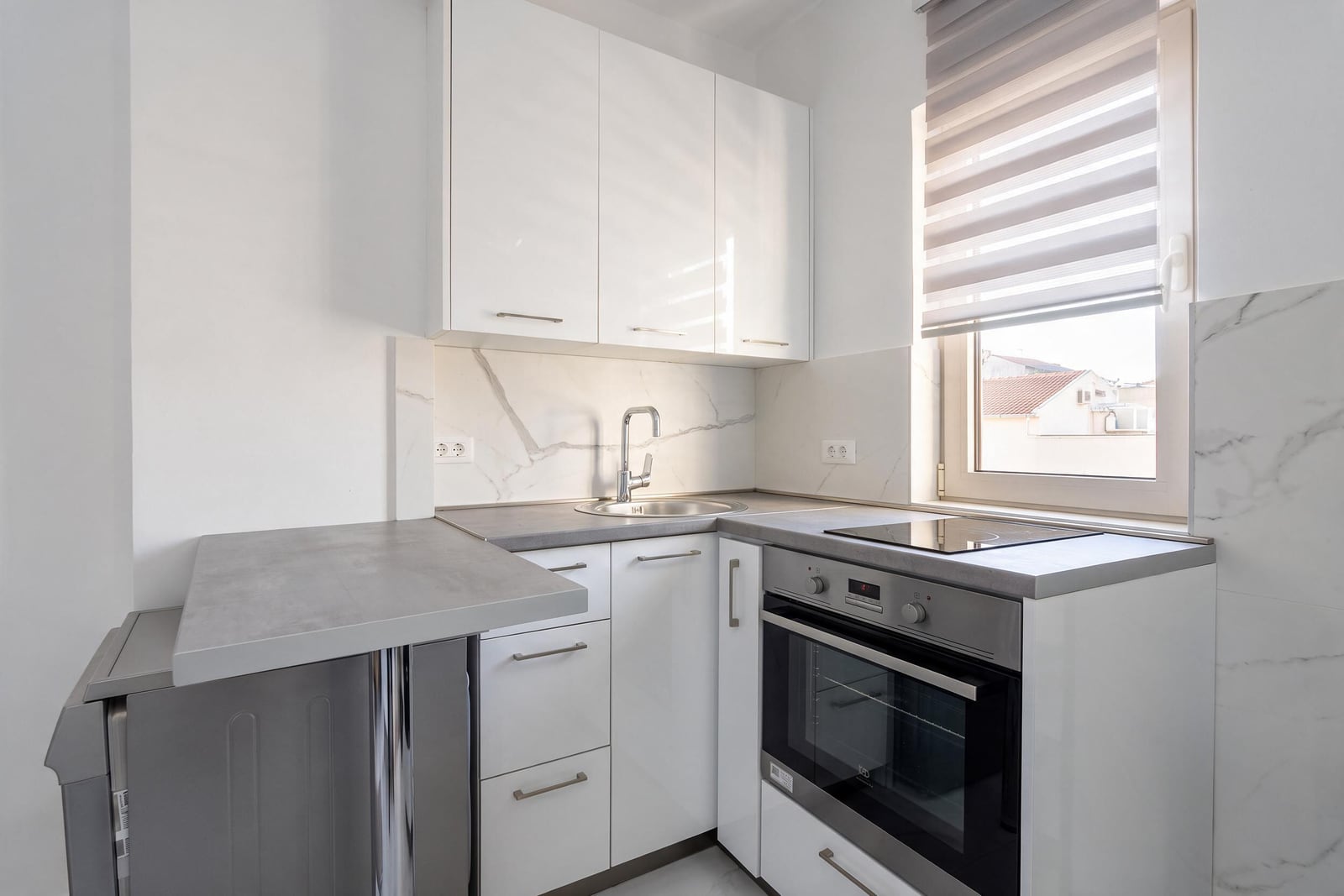 Modern kitchen with white glossy cabinets, grey countertop, built-in oven, induction cooktop, sink, and a window with striped blinds.