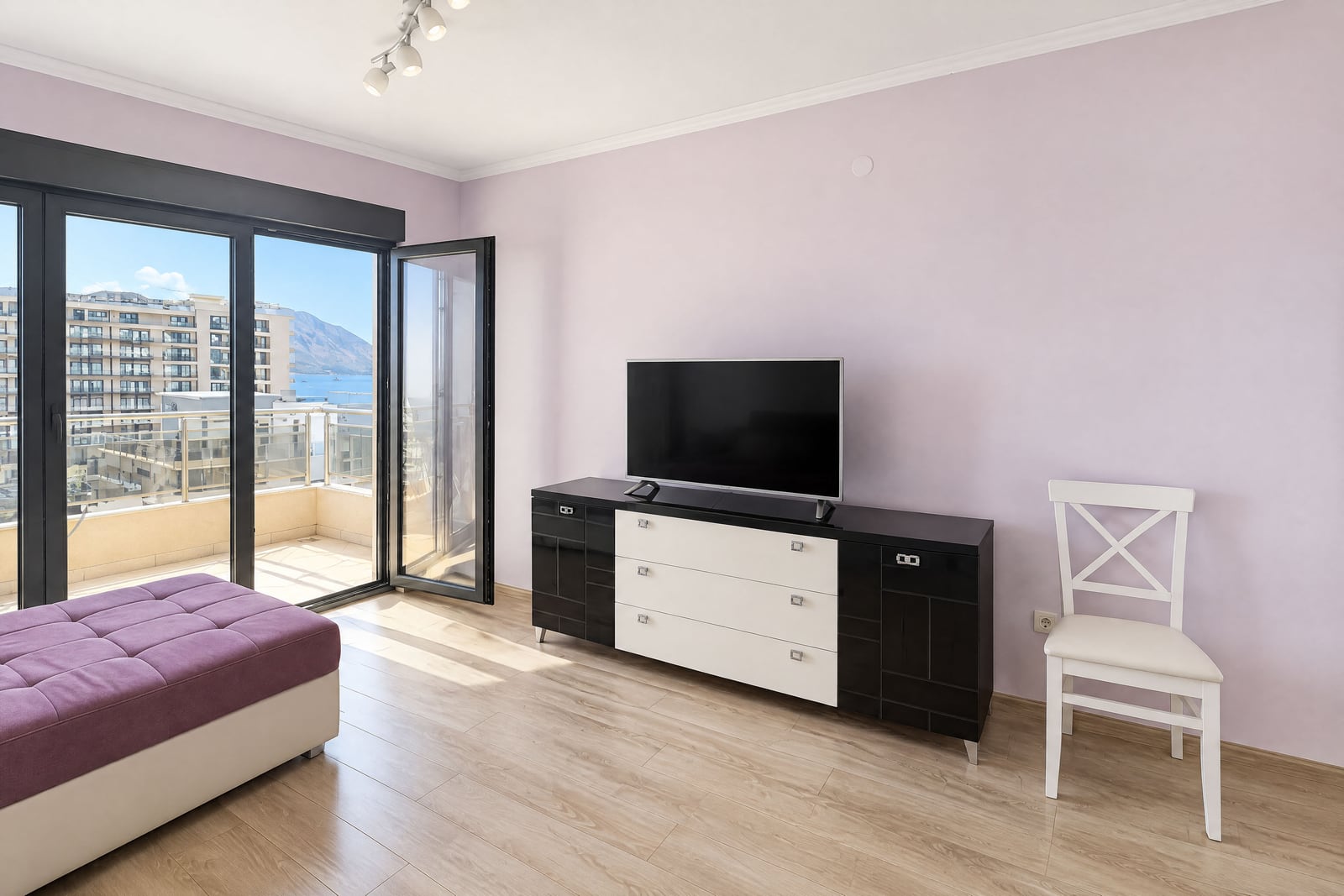 Living room with TV on a media console, purple ottoman, and open balcony doors overlooking the sea.