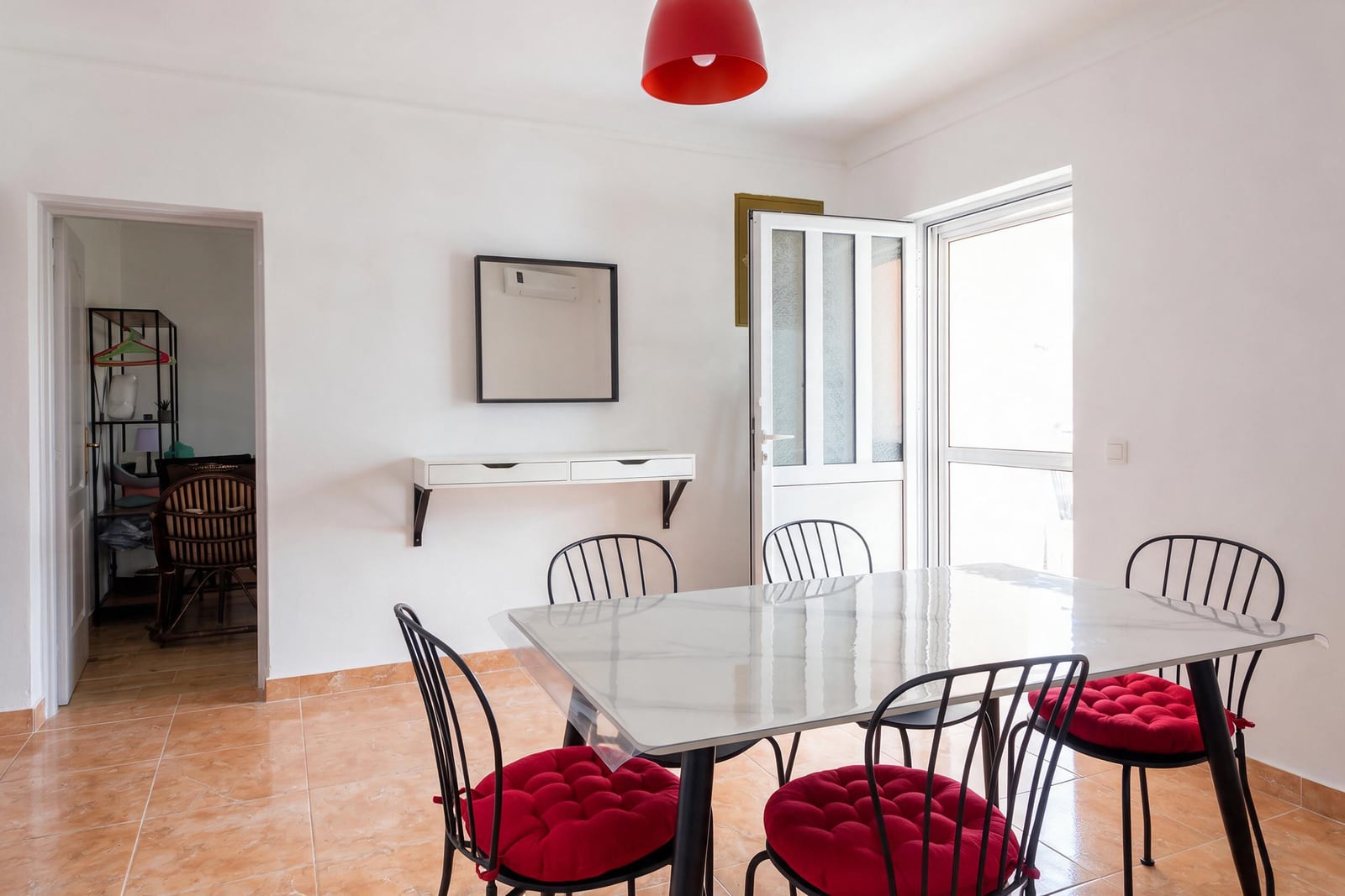 Dining area with a glass table, black chairs featuring red cushions, a wall-mounted shelf, and a red pendant light.