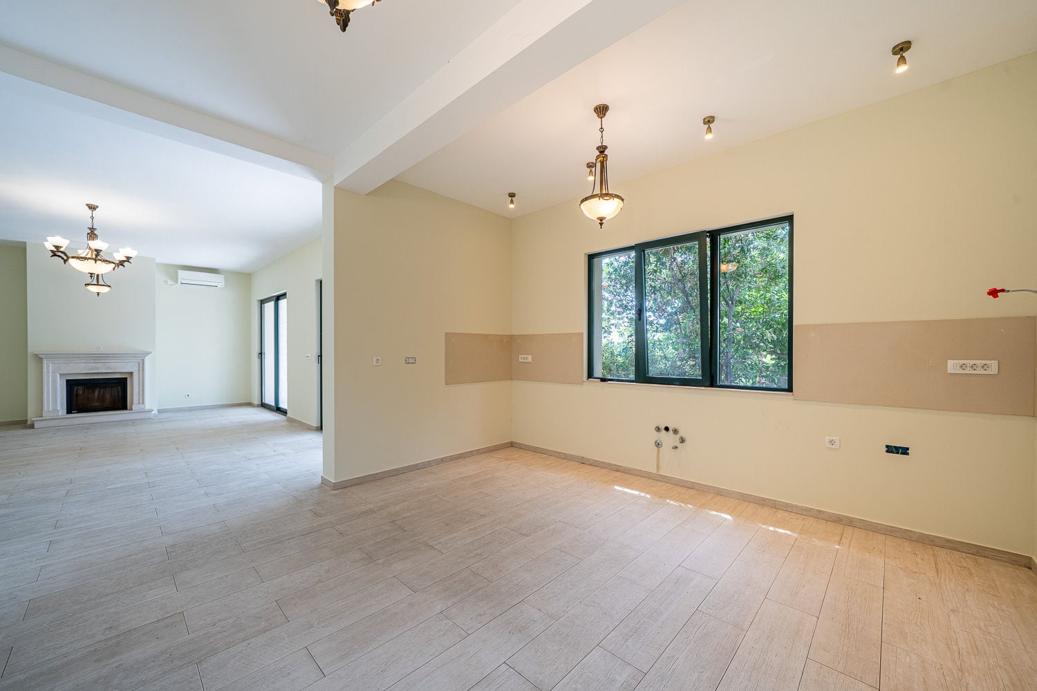 Empty room with a fireplace, chandeliers, light flooring, and a window overlooking trees.