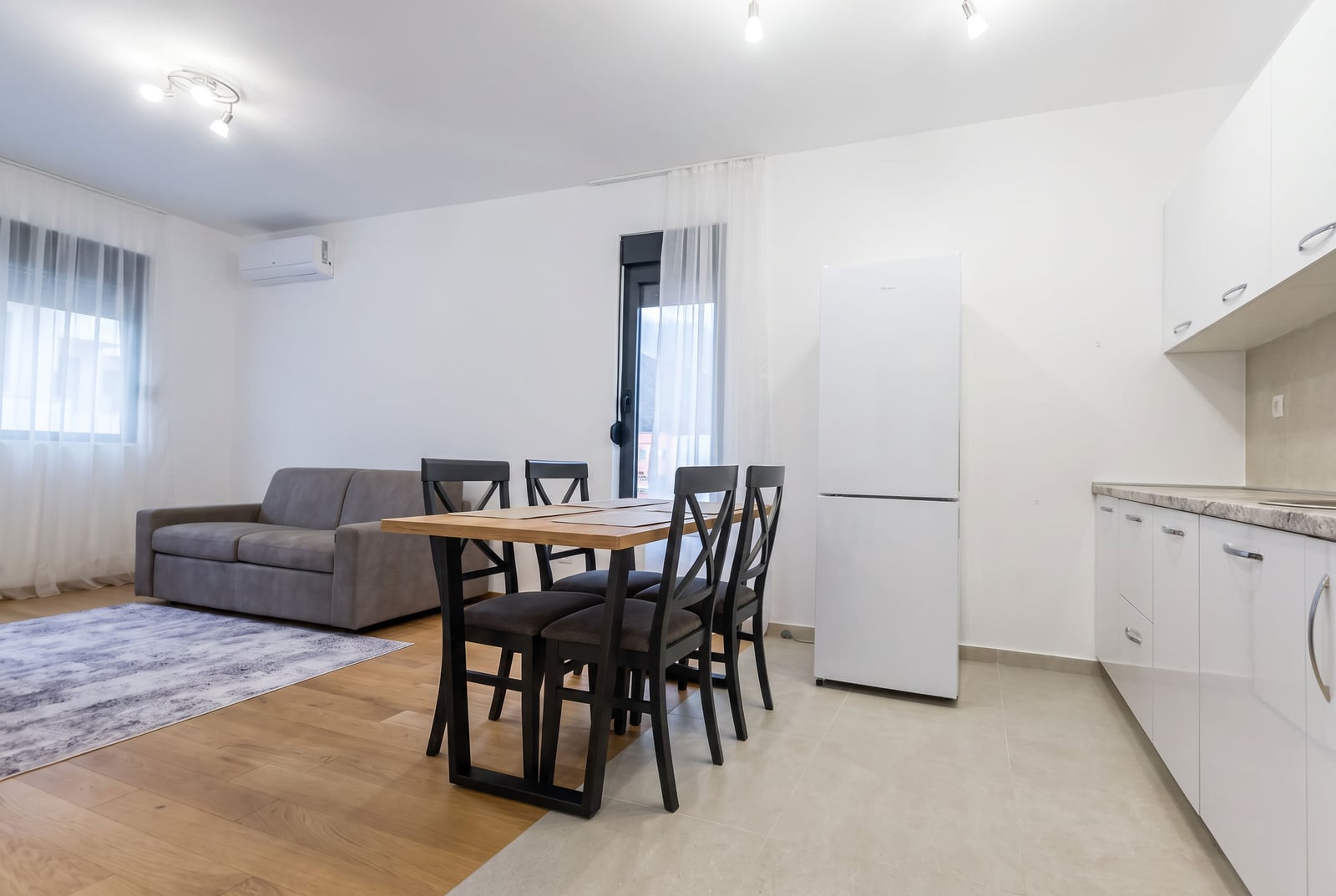 Living room with gray sofa, wooden dining table, black chairs, white kitchen cabinets, and refrigerator visible.