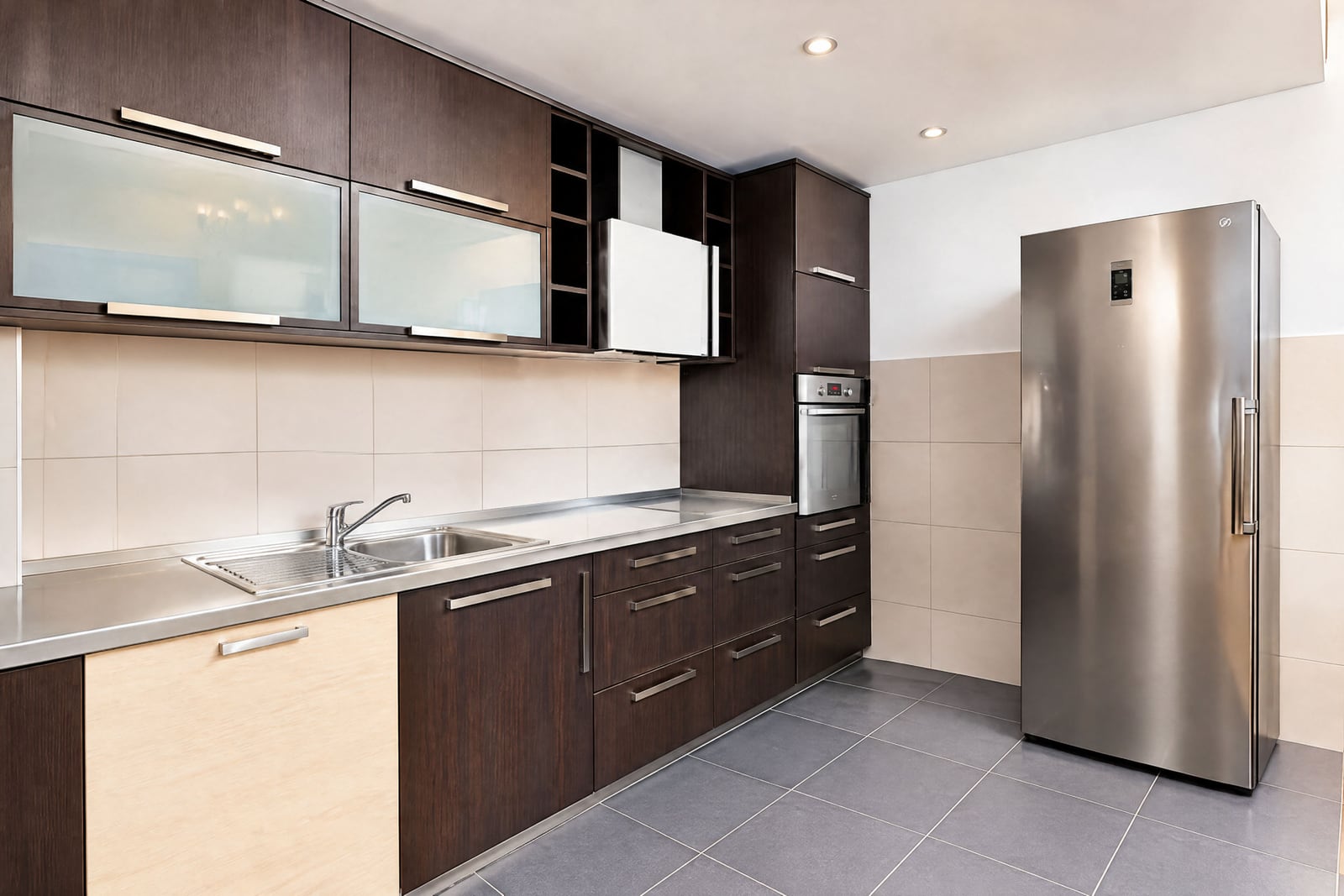 Modern kitchen featuring dark wood cabinets, stainless steel countertops, sink, built-in oven, and a tall refrigerator unit.