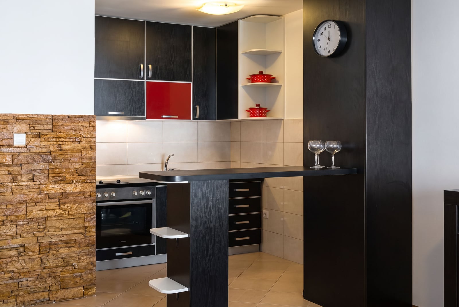 Modern kitchen with black and red cabinets, built-in oven, bar counter, stone wall accent, and tiled flooring.