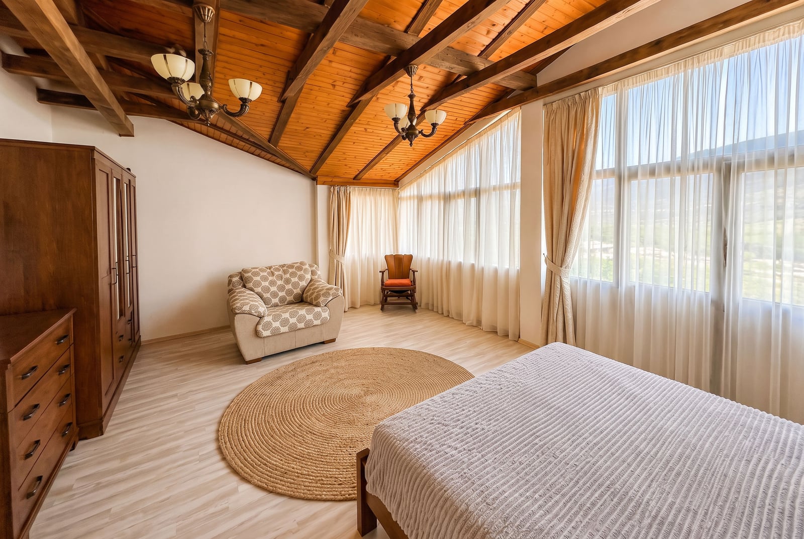 Bedroom with wooden beam ceiling, bed, wardrobe, armchair, and large windows.