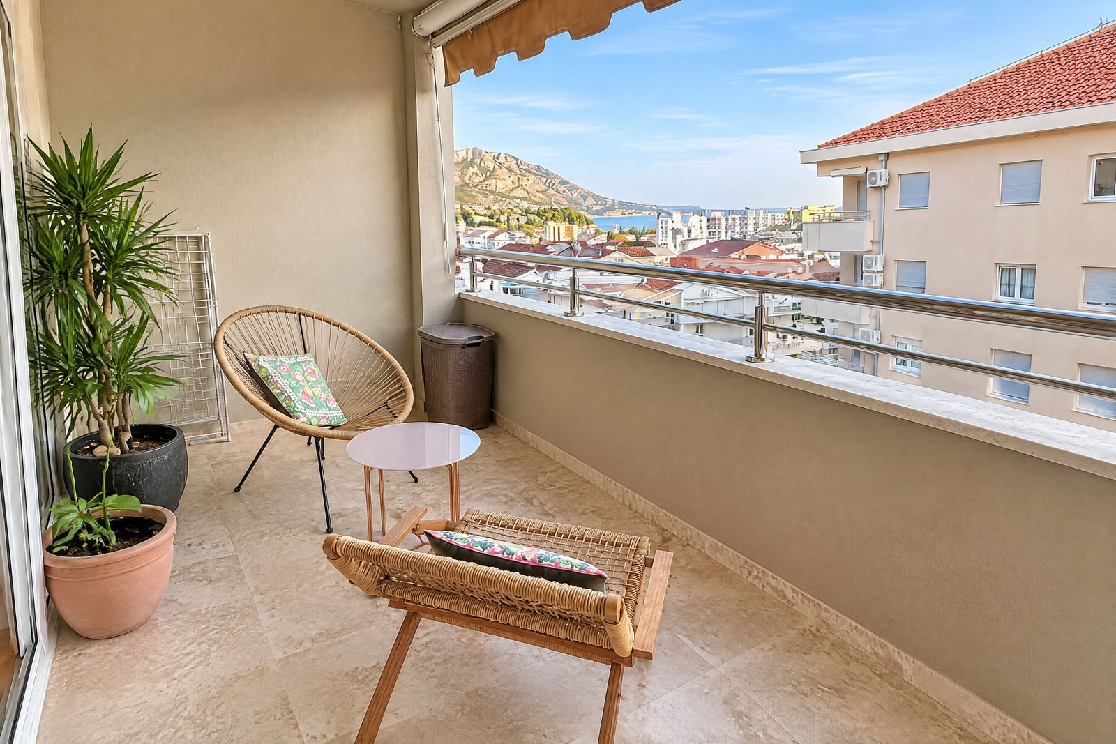 Balcony with wicker chairs, small table, potted plants, and a city-sea view.