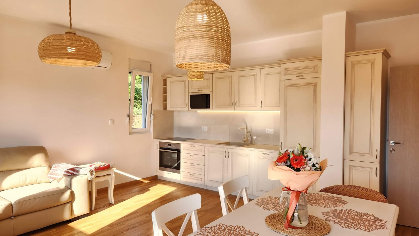 Bright living room with an open-plan kitchen in a Budva apartment, Montenegro: white furniture, wicker light fixtures, and a dining area.