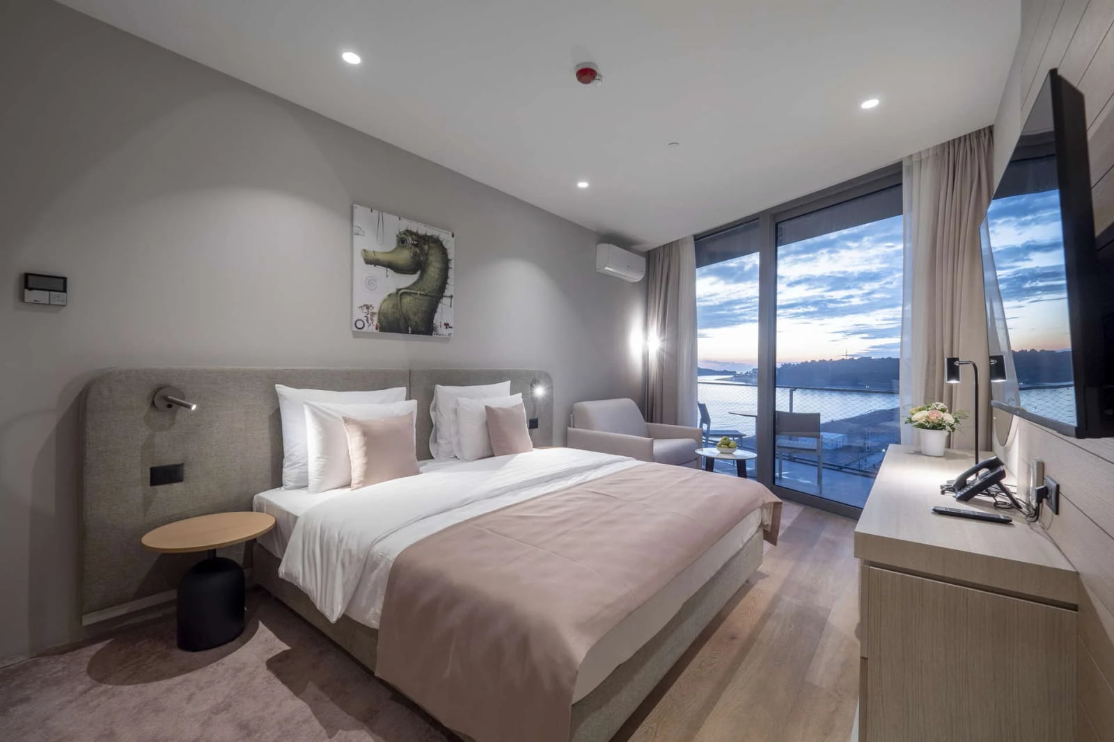 Modern bedroom featuring a large bed with white and pink bedding, a wooden dresser, TV, and floor-to-ceiling windows overlooking water.
