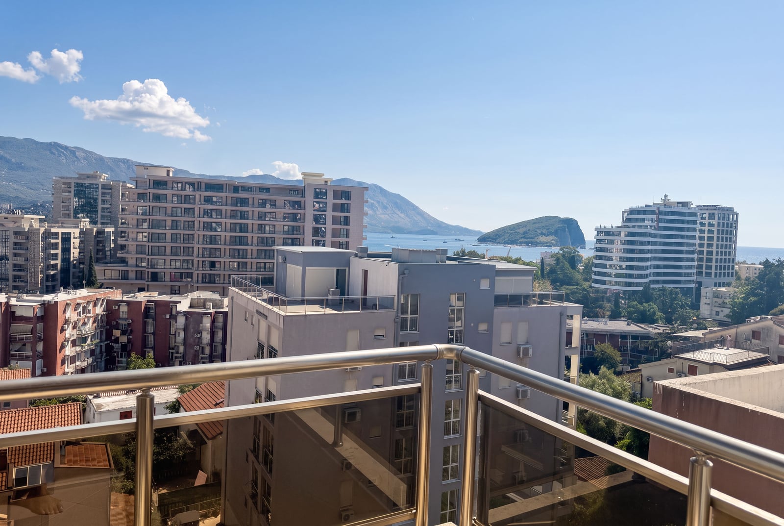 View from a balcony overlooking city buildings, sea, and mountains. Metal railing in foreground.