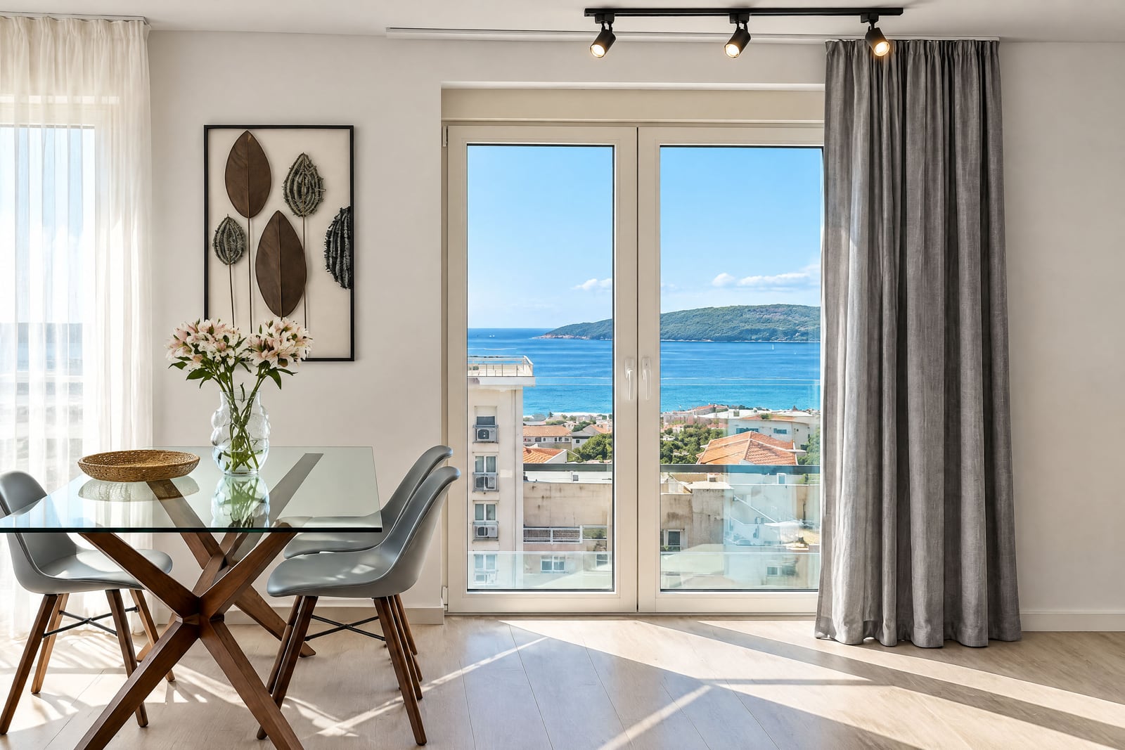 Bright room with a glass dining table, grey chairs, and a panoramic sea view through large balcony doors.